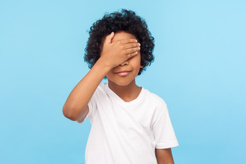 Fototapeta na wymiar I'm not watching. Portrait of cute little boy with curls covering eyes with hand not to look at something forbidden, hiding eyes playing peek a boo. indoor studio shot isolated on blue background
