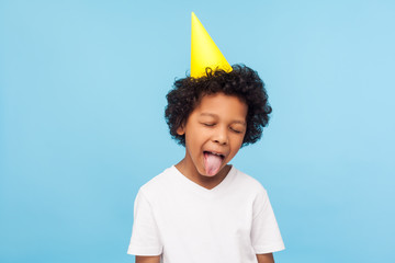 Portrait of adorable funny disobedient little boy with party cone on head sticking out tongue and...
