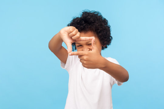 Portrait of curious nosy little boy in T-shirt looking through photo frame shape with fingers, focusing zooming at camera, viewing world with interest. indoor studio shot isolated on blue background
