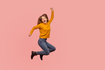 Vivid energetic ginger girl in sweater and denim jumping in air or flying up, shouting Yeah i did it, celebrating victory success, full of enthusiasm. indoor studio shot, pink background, full length