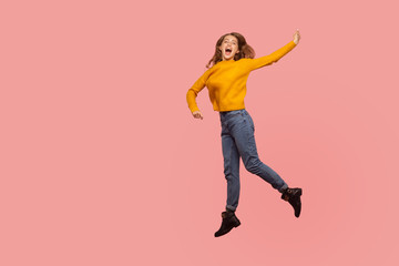 Fototapeta na wymiar Portrait of lively energetic ginger girl in sweater and denim full of enthusiasm jumping in air or flying up, screaming from happiness. indoor studio shot isolated on pink background, copy space