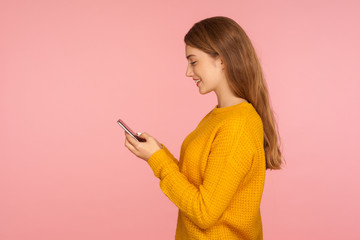 Side view of cheerful cute ginger girl typing message on cellphone and smiling, scrolling social...