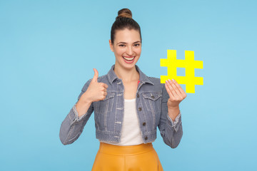 Like to popular blog and trends. Joyful attractive woman with hair bun in stylish outfit holding big yellow hashtag symbol and showing thumbs up gesture. indoor studio shot isolated on blue background