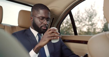 African American young businessman in glasses and tie sitting on the back sit of the car and sipping coffee early in the morning while tapping and surfing the smartphone.