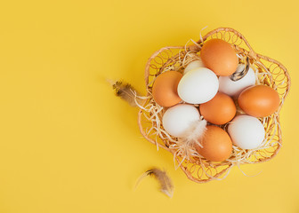 Easter concept. White and brown eggs with feathers in a wicker basket on a yellow background. Copy spase, top view