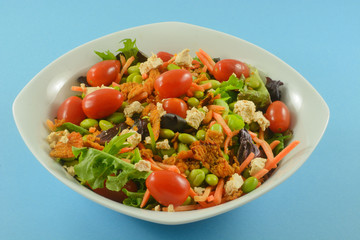 Healthy salad with fresh mixed greens, grape tomatoes, matchstick carrots, edamame, cheese and crispy red peppers in white bowl on blue background