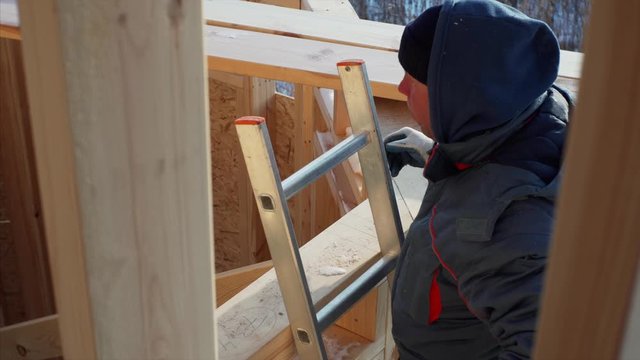 Builder in gloves is climbing up the steel ladder and measuring wooden plank on roof with measuring tape. Wooden frame house construction. Winter