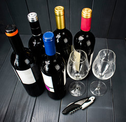 Bottles of red wine from different manufacturers with two sparkling wine glasses and a corkscrew for tasting, in well-lit gray wooden space