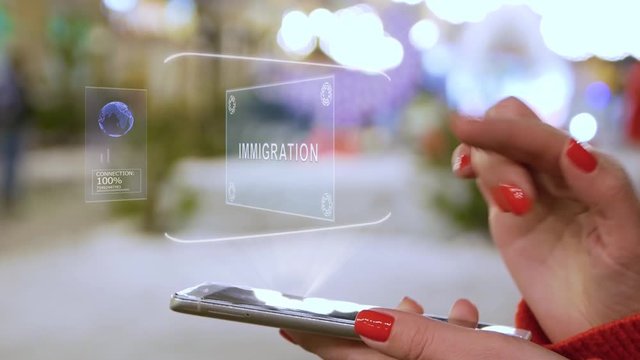 Female hands interact with HUD hologram text Immigration. Woman with red nails and sweater uses the holographic technology of the future in the smartphone screen on the background of street