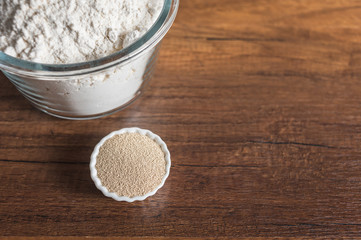 White flour in a glass bowl container and dry yeast in small ramekin on wood table surface.