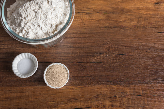 White Flour In A Glass Bowl Container, Dry Yeast, And Salt In Small Ramekin On Wood Table Surface.