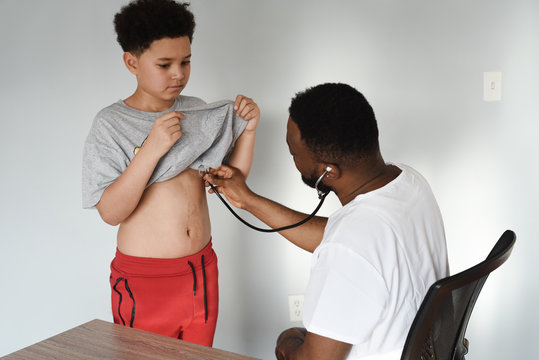 An African American Doctor With His Mixed Preteen Male. A Cute Black Paediatrician Examining His Young Mixed Preteen Boy Patient With A Stethoscope.