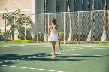 Tennis player woman playing on green hard court at resort hotel outside wearing white tennis dress...