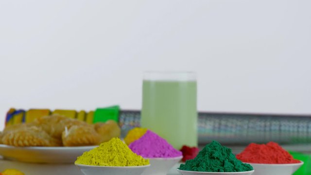Closeup shot of attractive festive items on the occasion of Holi the festival of colors. Bowls of organic Gulal colors rotating on a turntable with Kesar Thandai  yummy Gujiya  and designer water g...