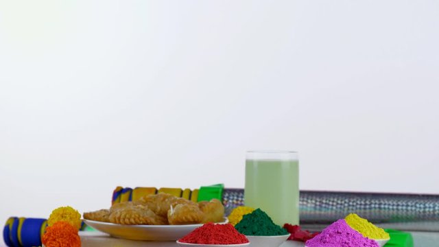 Pan shot of beautifully placed festive items for the joyful Indian festival - Holi. Containers of colorful Gulal rotating on a turntable with Kesar Thandai  Gujiyas  and designer water guns (Pichka...