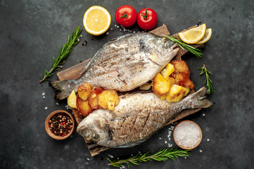 grilled dorado fish and fried potatoes served on a cutting board with spices and lemon on a stone background