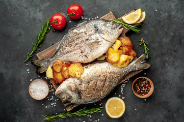 grilled dorado fish and fried potatoes served on a cutting board with spices and lemon on a stone background