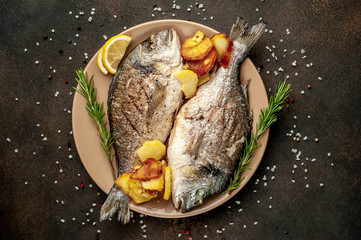 grilled dorado fish and fried potatoes on a plate with spices and lemon on a stone background
