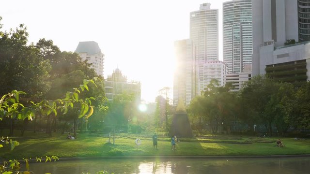 People Relax in Public Park with Lake and Green Trees with Skyscrapers Buildings in the City on Background and Bright Sunshine of Sunset. Park for Rest, Leisure and Fun in Center of Modern Metropolis
