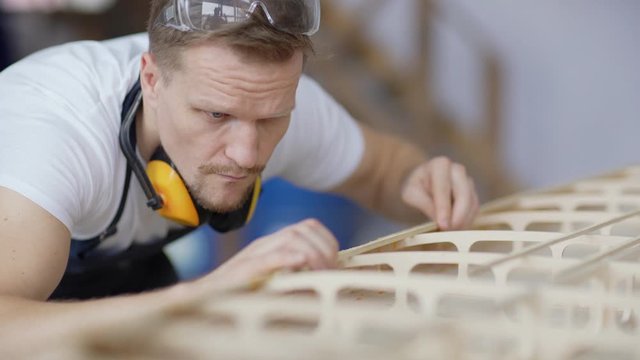 Closeup tracking shot of focused middle aged carpenter constructing wooden boat model placed on workbench in woodworking shop