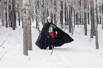 Medieval warrior in armor, helmet, black cloak with a saber in his hands in the winter in the forest.