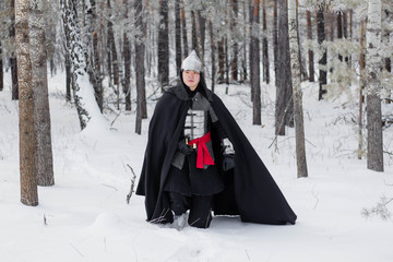 Fototapeta na wymiar Medieval warrior in armor, helmet, black cloak with a saber in his hands in the winter in the forest.