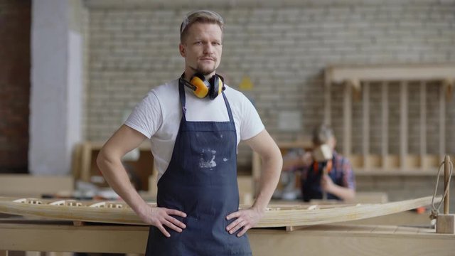 Portrait video of middle aged carpenter in apron looking at camera posing with his hands on hips while his little son learning to woodwork in background in carpentry shop