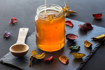 Honey jar still life on black slate background with dried flowers scattered and wooden spoon -...