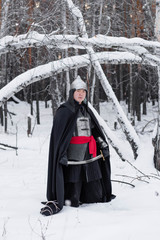 Medieval warrior in chain mail, helmet, black cloak with a saber in his hands against the background of a winter birch forest.