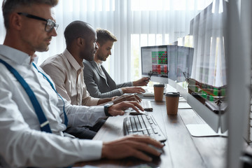 Try and try, earn and learn. Three businessmen sitting at desktop front PC computers with financial graphs and statistics on monitors. Analysis of digital market and investment. Stock trade