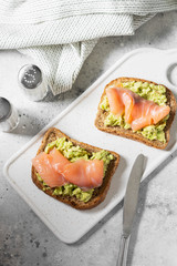 Bruschetta with avocado and salmon on a white ceramic plate on a light gray kitchen table. Toasted bread with avocado and red fish. The concept is helpful and a delicious Breakfast
