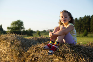 A romantic carefree little girl sits on a haystack in a field against the background of a forest, enjoying the freedom and tranquility of rural nature during the holidays. Incredible colorful sunset