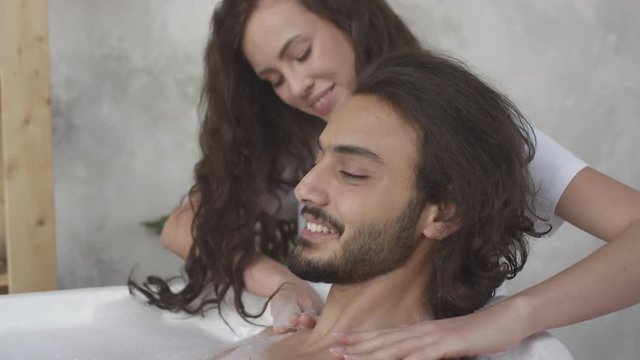 Arc shot of romantic young woman smiling and massaging shoulders of her handsome boyfriend while he lying in bath
