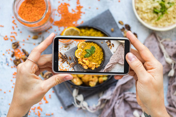 Woman take picture of vegan food with phone at her kitchen. Hand make a closeup smartphone photo of Indian masala lentil for blogging or social media content. Vegetarian healthy food.