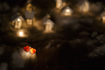 Two red glass hearts on the snow in front of a garland of wooden houses with a light in the window. Symbols of love on Valentine's Day and home warmth and comfort, background with a warm snow village.