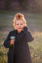 a curly haired blonde girl with a tuft of hair in the background light blows a dandelion in a coat in a meadow in the autumn