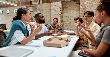 Happy team relaxing at work. Positive multicultural team eating pizza and communicating with each...