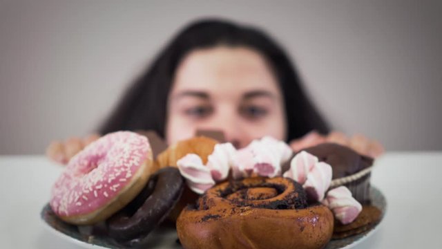Plate with sweets standing on the table, obese Caucasian woman appearing from under the table and smiling. Hungry fat girl looking greedily at dessert. Obesity, overweight problem, unhealthy eating.