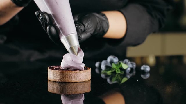 Pastry chef decorates biscuit with purple cream from pastry bag, close-up. Preparation of blueberry cake at commercial bakery with piping bag