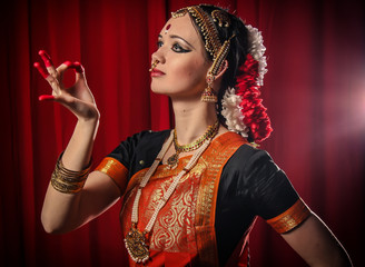 Portrait of white girl as an Indian classical dancer in traditional dress and performing dance performance on the red curtain background. Classical indian temple dance form Bharatanatyam. Dance pose