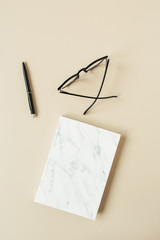 Marble styled notebook, pen, glasses on pastel beige background. Minimalist home office desk table. Flat lay, top view education, work composition for blog, social media.