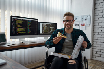Pizza time. Portrait of cheerful male web developer in a wheelchair eating pizza and smiling while...