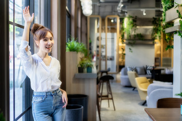 Asian cheerful woman waiting friend at coworking space, raise hand up to say hello her coworker with smiling face and happiness to meet. Freeland working outside office for inspecting a customer area