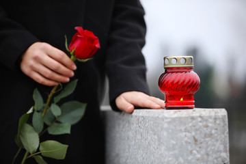 Woman holding red rose near grey granite tombstone with candle outdoors, closeup. Funeral ceremony