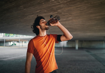 Young male athlete with earphone in his ears drinking water from reusable bottle standing under the...
