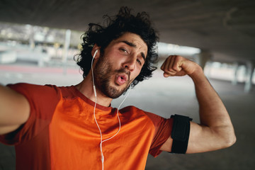 Attractive fit young sportsman with earphone making funny face flexing his muscles while taking a...