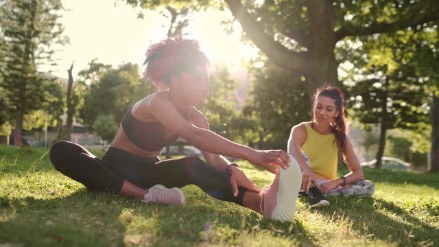 Smiling sporty young woman in sportswear stretching their legs while sitting on green grass with her friend in the park on sunny day