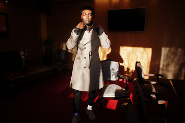 Handsome african american man posing  inside room with sunlight shadows in black hat and beige coat.