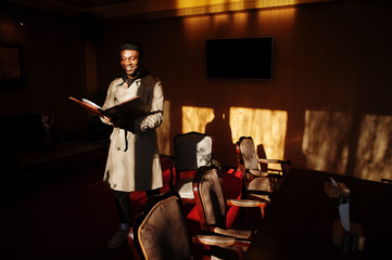 Handsome african american man posing  inside room with sunlight shadows in black hat and beige coat.