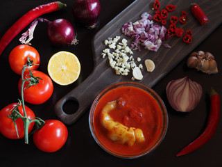 Traditional soup puree, tomato sauce salsa in a glass bowl, a branch with tomatoes, hot pepper, garlic, onion, linen napkin on a black background closeup. Vegetable dish recipe for proper nutrition.